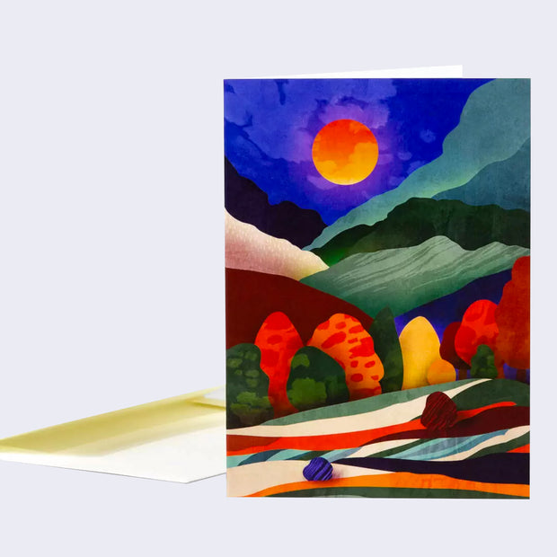 Notecard with a full bleed illustration of a night scene, made primarily out of cut out shaped and simplistic outlines. Scene is a night sky with a bright orange moon over many mountains and trees.