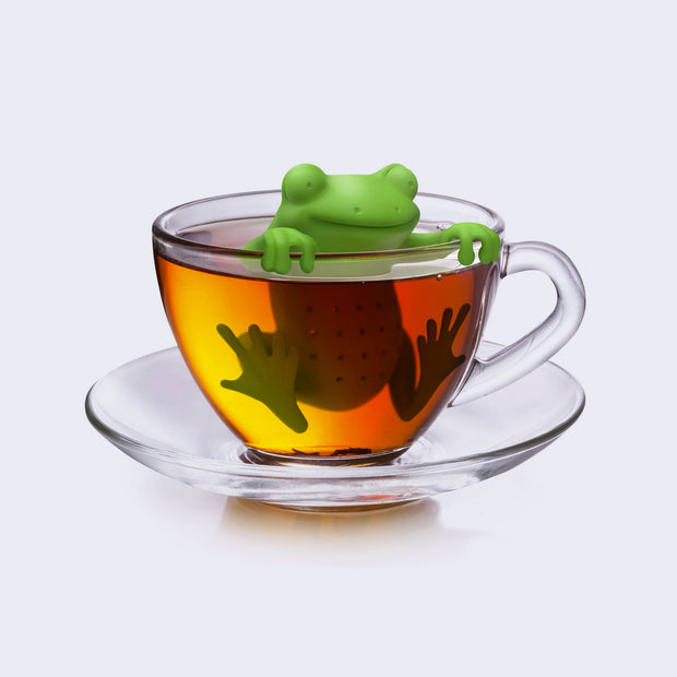 A silicone green frog shaped tea infuser inside of a clear mug with tea inside of it. Frog is smiling and holding itself up with its hands while its body is submerged in the tea, its feet up against the inside of the mug.