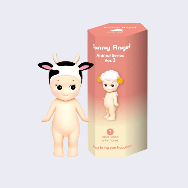 Vinyl figure of a Kewpie baby standing nude with a cow patterned hood hat atop its head. It stands next to a pink display box that reads "Sonny Angel Animal Series Ver. 2"