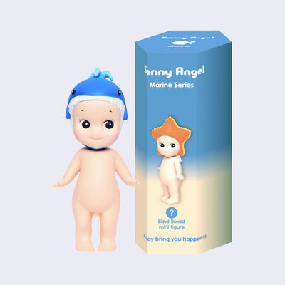 Vinyl Kewpie baby figure, standing nude while wearing a whale shaped hat on its head. Beside is its display box, blue to cream ombre with a small Kewpie baby wearing a starfish shaped hat.