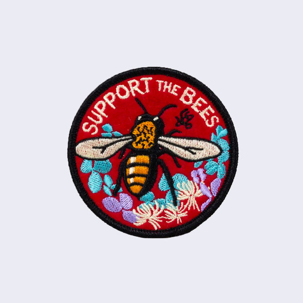 Round red patch with a black outline of a bee, flying over some abstract flowers and leaves. "Support the Bees" is written along the top.