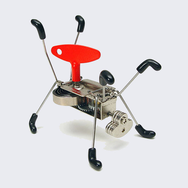 Small robotic toy with a rectangular body, 8 thin wire legs with black boots on each. A large red crank is attached to its main body and it has a small circle for a head.